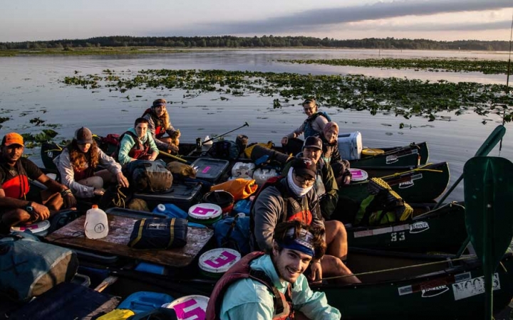 A group of people sitting in canoes that are tied together smile at the camera. They appear to be floating in shallow water. 